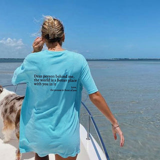 Turquoise t-shirt with inspirational message "Dear person behind me, the world is a better place with you in it" worn by a person overlooking the water.