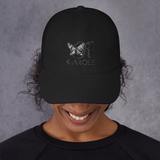 Black dad hat with K-AROLE logo in a minimalist style, worn by a smiling person against a plain gray background.