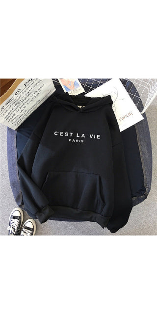 Casual black hoodie with "C'est la vie Paris" text graphic, sitting on a surface with papers and other accessories in the background.