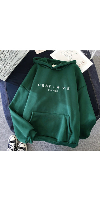 Casual green hoodie with 'C'est la vie Paris' text printed on the front, displayed on a white background along with a pair of black sneakers.