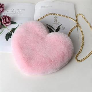 Plush pink heart-shaped shoulder bag with a gold chain strap, resting on a floral book