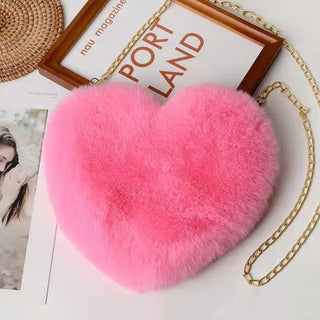Soft, plush heart-shaped shoulder bag in vibrant pink color with chain strap, displayed on a wooden frame with magazine on a round woven mat.