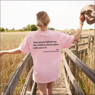 Attractive young woman wearing a pink t-shirt with an inspirational slogan printed on the back, posing on a wooden footbridge in a grassy field.