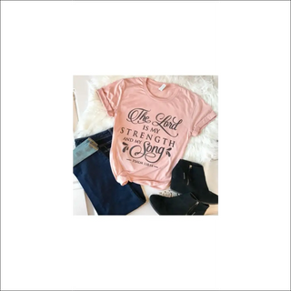 Pink women's fashion T-shirt with "The Lord is my strength and my song" slogan graphic, laid on a white background with a furry blanket or rug.